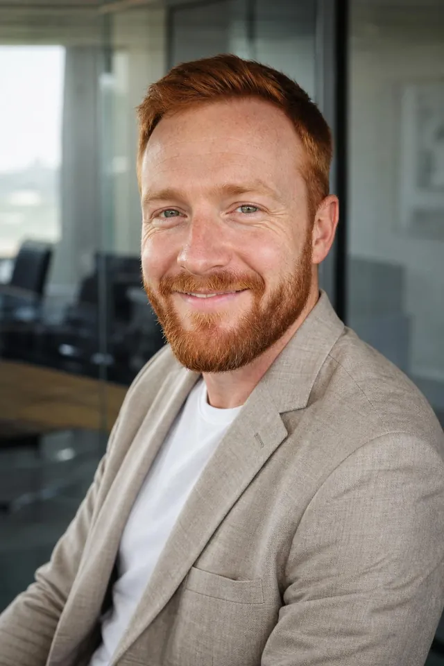 Man with a red beard in a beige blazer over a white tee in a glass-walled office
